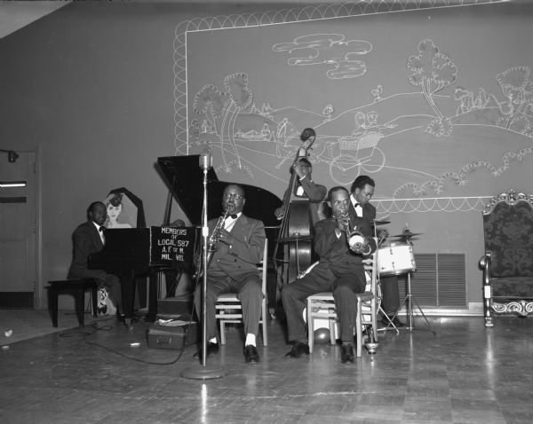 The Norman Ebron Band, members of the American Federation of Musicians, segregated Local 587, play coronet, saxophone, piano, drums and standup bass in a dance hall. A large line-drawing painting of a drive in the country decorates the wall behind the band.