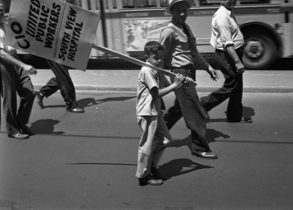 Close-up of two participants, a man and a boy, in a United Public Workers Parade/Demonstration. Both are looking at the photographer and the boy carries a sign that reads "CIO United Public Workers South View Hospital." In the background a few other participants are walking along and a bus is driving by.