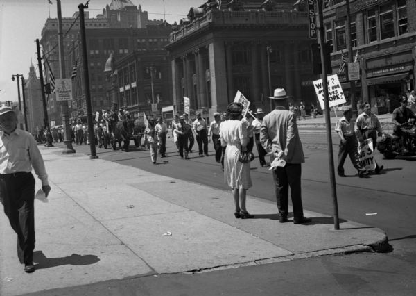 A United Public Workers parade/demonstration from an adjacent sidewalk. The line of participants including a horse-drawn wagon stretches into the distance. Signs read "Big Three Unity" and "Why Must Our Wives Work?" Large downtown buildings line the opposite side of the street.