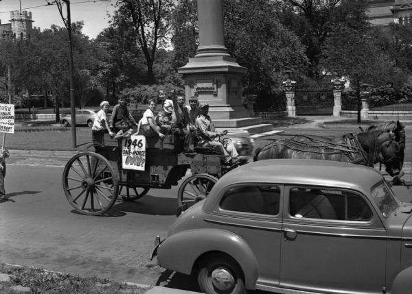 View from sidewalk of horse-drawn wagon on a tree-lined city street in a United Public Workers parade/demonstration. The wagon has a number of boys in it who are looking at the photographer and displays a sign that reads "1946 One Horse Pay!" A parked car is in the foreground and a stone monument is in the background on a grassy median.