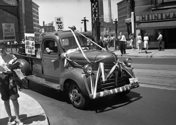 Decorated truck driven by Leonard Greachowiak through downtown in a United Public Workers Parade/Demonstration. The beribboned pickup displays a sign that reads "Butter 81¢ .lb Steak $1.45 .lb". A few pedestrians stand on the sidewalks.