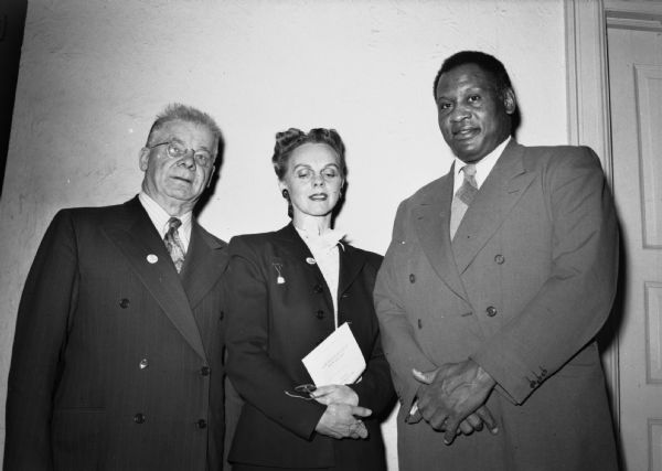 Paul Robeson, Mrs. Emil H. Jones and Leo Krzycki pose for a group portrait at the Win the Peace Luncheon. They are wearing suits and standing in front of a blank wall.