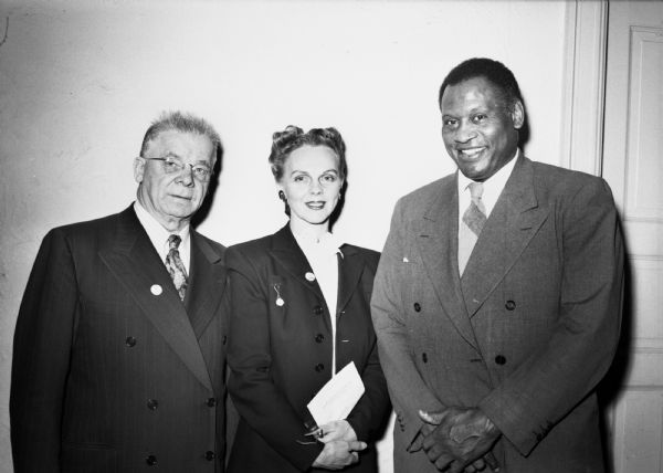 Paul Robeson, Mrs. Emil H. Jones and Leo Krzycki pose for a group portrait at a Win the Peace Luncheon. They are wearing suits and standing in front of a blank wall.
