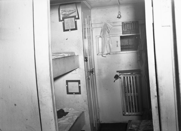 Worker's bunks on a railroad car ferry. The spare room has a calendar, light, radiator, table, locking closet and two bunks. Workers' clothes hand on the wall and a picture is tucked behind what looks like some kind of official posting. Part of a series on working conditions aboard Lake Michigan railroad car ferries. Ferry workers belonged to the National Maritime Union (NMU).