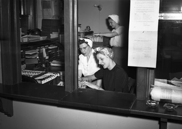 View through a reception window of the three women union office staff of UAW local 75 (Seaman Body Plant) looking at files and cards. They are Audrey Wergin, Leona Hesse and Claudia Keim. The women have their hair up in kerchiefs, and a large stocked desk is visible in the background.