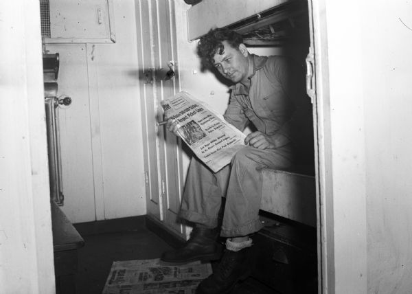 Worker reads union newspaper while sitting on a bunk and smoking a cigar. Part of a series on working conditions aboard Lake Michigan railroad car ferries. Ferry workers belonged to the National Maritime Union (NMU).