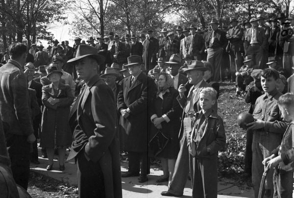 Outdoor rally of Allis-Chalmers workers. A man is speaking into a microphone in the front and people stand on the sidewalk and the grassy rise in the background. Trees encircle the natural amphitheater.