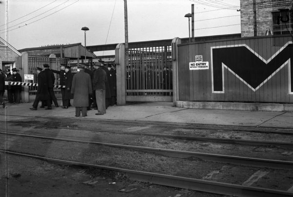 Allis-Chalmers factory gate during strike. Railroad tracks are in the foreground and a large group of police officers stand inside of and coming out of the gate.