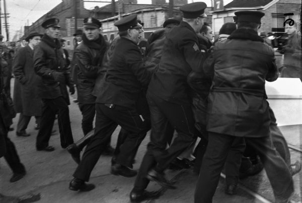 Police pushing a group of strikers in front of the factory gate at the Allis-Chalmers strike. On the far right is a man holding a press camera.
