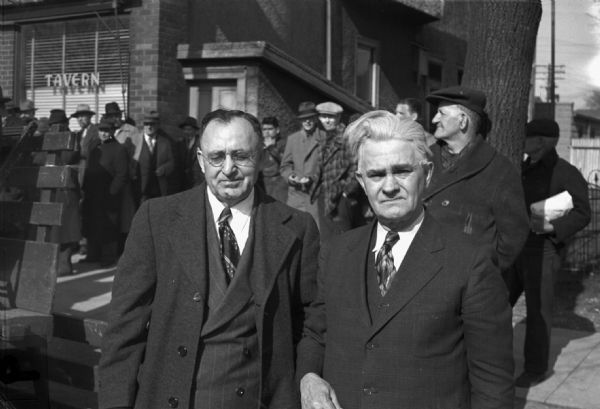 Mayor Vincent Totka of Cudahy and Mayor Charles Plotz of South Milwaukee pose for a photograph after greeting a CIO organized rally in South Milwaukee. A line of men stand behind them on the sidewalk in front of a tavern.