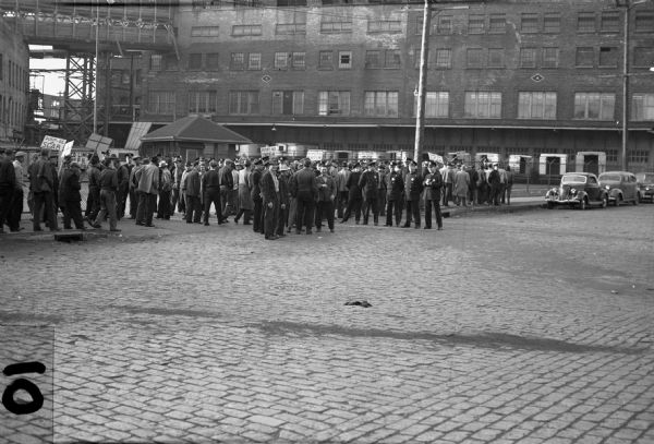 Striking workers and police outside of Plankinton Meat Packing.  Strikers are standing on the cobblestone street and sidewalk holding signs that read "Don't Be A Scab," "9 Cents Not Enough!" and "Respect Our Picket Line."