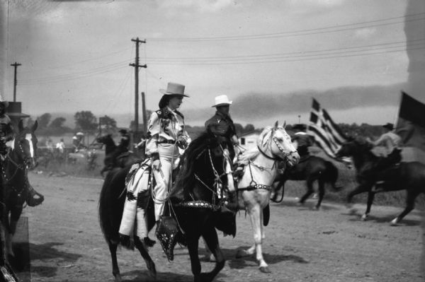 Cowgirl and cowboys riding horses at the Cedarburg Horse Show. In the background are two more men on horses carrying flags.