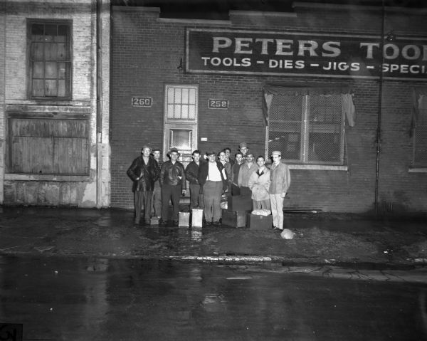View from across street of employees of Peters Tools, Local 100, standing on the sidewalk in front of the brick storefront with a number of cases at their feet. The sign, which is partially cut off, reads "Peters Tools: Tools-Dies-Jigs-Specia..."