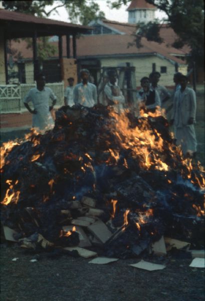 Bank burning money in Tezpur, Assam during the Sino-Indian Border Conflict. The large pile of paper is steadily burning and a group of men are watching from behind. There are large buildings in the background.