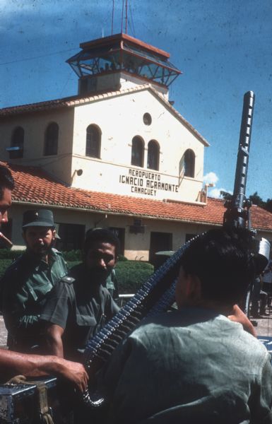 A group of soldiers, including Juan Almeida Bosque (center), operating chief of the Cuban Air Force, are standing outside of Camaguey Airport, Cuba. The soldiers are wearing fatigues, and one soldier is holding a large automatic gun and a bandoleer of bullets. The cream and terracotta colored airport building and control tower are in the background. The sign reads: "Reropuerto Ignacio Agramonte Camaguey."