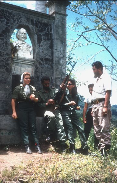 Juan Almeida Bosque, operating chief of the Cuban airforce, and a small group having a picnic in the countryside of Cuba. Three soldiers in fatigues are sitting on an old stone wall with an inset bust. Bosque is holding a gun. A man and a boy are standing on the right, and green rolling mountains stretch into the distance behind them.
