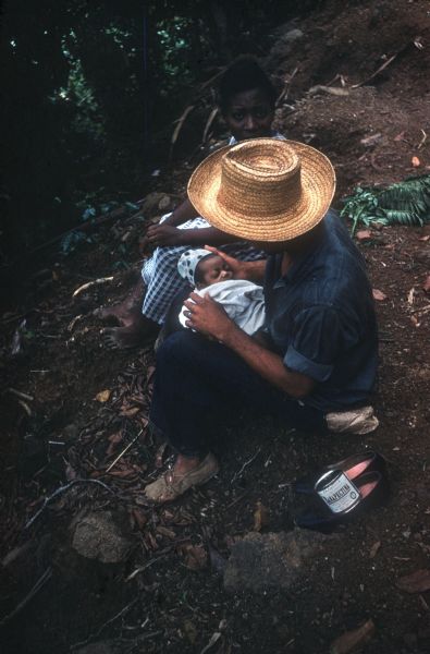 A man wearing a straw hat is sitting on the ground holding a baby in the countryside in Cuba. A woman sitting behind him is looking at the photographer. The woman's shoes are sitting in the foreground with a can of food.