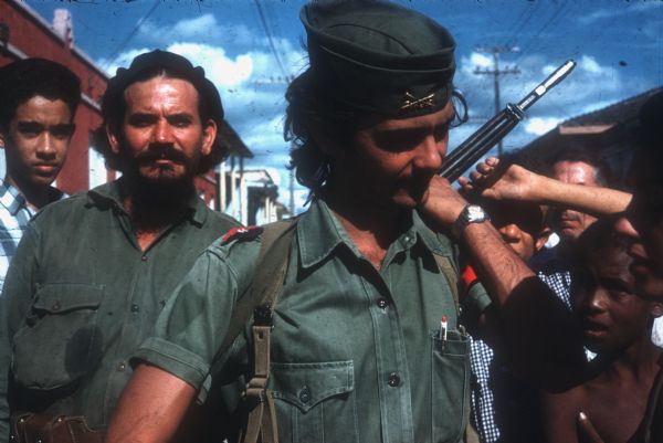 Close-up of two soldiers walking down the street in La Maya Cuba one year after the Cuban Revolution. Boys are crowding around the soldiers, and town buildings are in the background. Photograph revisits the site of a battle Chapelle photographed the year before.