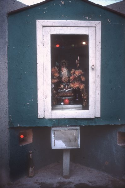 Shrine for police officers in Cuba one year after the Cuban Revolution. The shrine is a blue plaster enclosure with a white-framed glass door.  Inside is a figurine of the Virgin Mary and child surrounded by fake flowers. Colored lights illuminate the shrine. There is an unopened bottle of what is probably beer on the ground below the shrine.