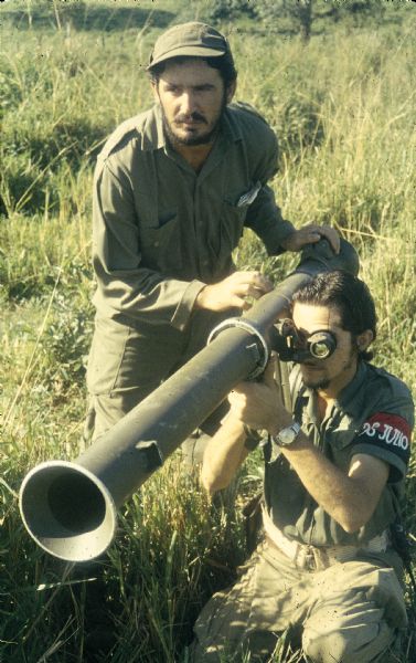 Fidel and Raúl Castro pose with a bazooka for a photograph in a field during the Cuban Revolution. Fidel is standing above on the left and Raul is kneeling with the weapon and looking through its scope.