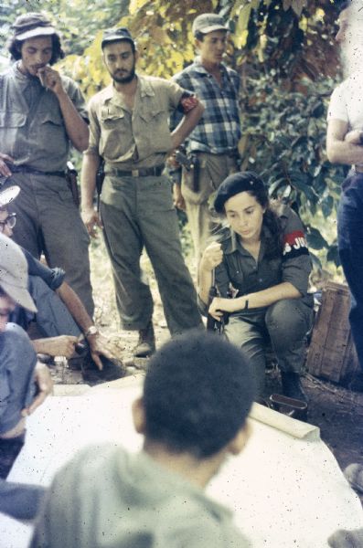 Meeting of the 26th of July Movement in the woods of Oriente Province during the Cuban Revolution. A number of participants, including Vilma Espin and Juan Almeida Bosque, are sitting, standing and crouching around a map laid on the ground in the country.