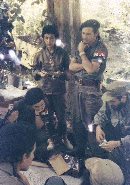 Meeting of the 26th of July Movement in Oriente Province during the Cuban Revolution.  Vilma Espin and Fidel Castro crouch over a map spread on the ground.  Celia Sánchez and Raúl Castro stand behind them next to a tree.