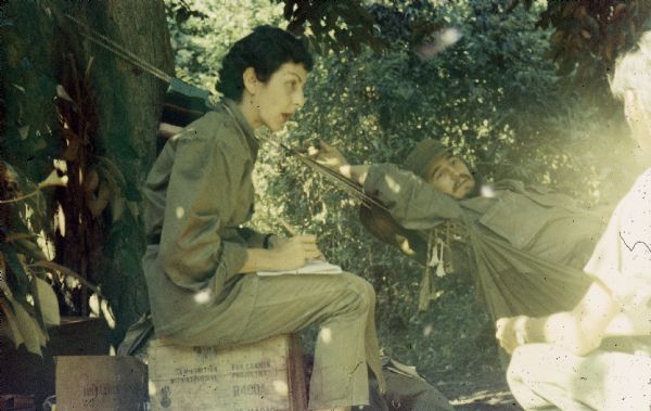 Informal meeting in the woods in Oriente Province during the Cuban Revolution. An unidentified man is crouching in the foreground, Fidel Castro reclines on a hammock and Celia Sánchez is sitting on a wooden crate writing.