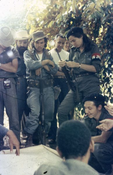 Meeting of the 26th of July Movement in Oriente Province during the Cuban Revolution. Vilma Espin stands in the middle with a gun, and Raúl Castro and Juan Almeida Bosque crouch next to a map spread out on the ground in the countryside. Other movement participants stand in a row in the background.