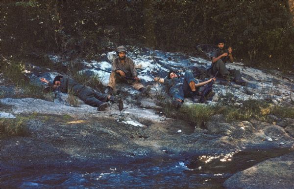 26th of July Movement soldiers resting by a stream in Oriente Province during the Cuban Revolution. Two soldiers aresleeping on the sandy bank and two soldiers are sitting up with their rifles at the ready.