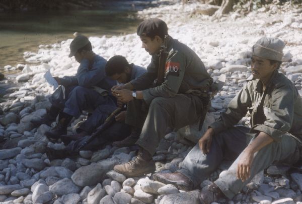 26th of July Movement soldiers rest by a river in Oriente Province while on patrol during the Cuban Revolution. They are sitting on a rocky bank of the river. One soldier is reading, another is cleaning his gun and the other two are sitting thoughtfully.