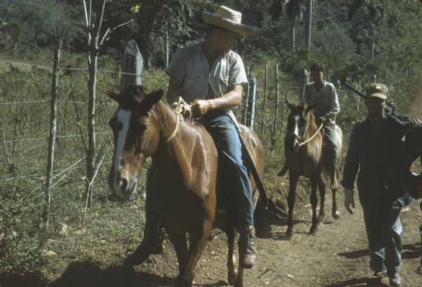 26th of July Movement soldiers on patrol in Oriente Province during the Cuban Revolution. One soldier is walking along a dirt road carrying a large automatic gun over his soldier. Two boys on horses with rope bridles are accompanying the soldiers down the dirt road bordered by a barbed wire fence.