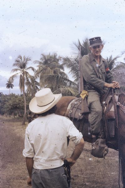 26th of July Movement scout on horseback in the countryside in Oriente Province during the Cuban Revolution. The scout is talking to a man who is standing on the ground, and there is a row of palm trees in the background.