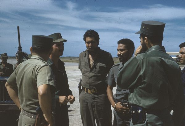 Group of soldiers talking on a runway at the Camaguey Airport in Cuba one year after the Cuban Revolution. Juan Almeida Bosque, the operating chief of the Cuban Air Force, is standing second from right, and a military jeep with a large gun is parked in the background.