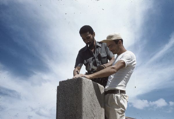 Juan Almeida Bosque, operating chief of the Cuban Air Force, hammering a spike into a block of concrete with the help of a construction worker.
