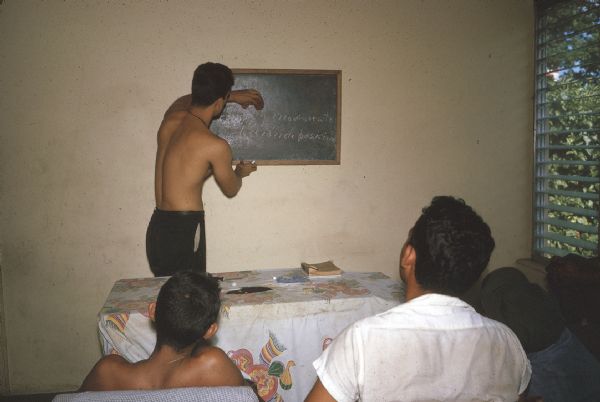 A meeting of the anti-Castro group Commandos L. A man is standing at the front of the room writing on a chalkboard and other members are sitting and listening.