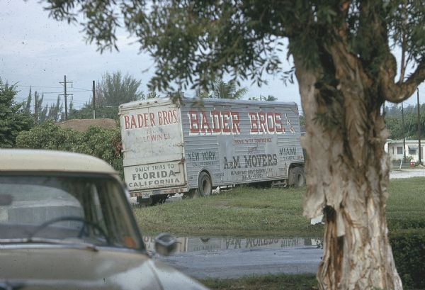 Moving truck across the street from the house of the anti-Castro group, Commandos L, purportedly occupied by CIA observers of the group. The moving van reads "Bader Bros. Inc." and "Daily Trips to Florida California" and is parked on a lawn in a suburban neighborhood.