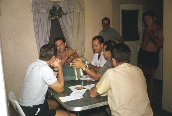 Meeting of the anti-Castro group Commandos L. Men are sitting and standing around a kitchen table with a radio receiver and transmitter. Tony Cuesta is sitting at one end of the table in a red plaid shirt.