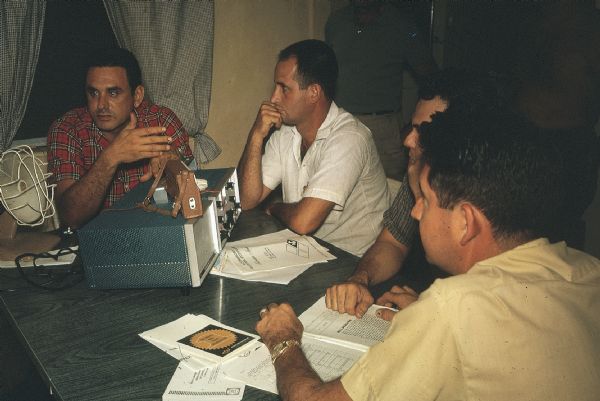 Meeting of the anti-Castro group Commandos L with radio transmitter and receiver. Four men are sitting around a kitchen table with a radio transmitter and receiver on it looking at manuals and instructions. A fan is on the table and Tony Cuesta is sitting next to it.