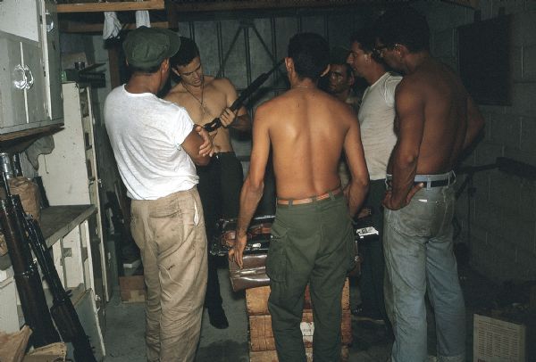 Members of the anti-Castro group Commandos L in a garage functioning as an improvised armory. Group members gather around a stack of crates and are watching a man handle a shotgun.