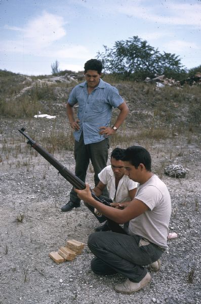 Members of the anti-Castro group Commandos L practicing rifle marksmanship in the Everglades. A man with a rifle is crouching on the ground next to boxes of ammo, and two other men are looking on in an open gravelly area.