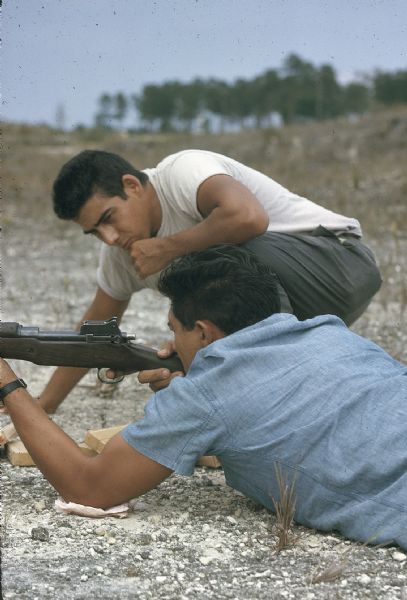 Two members of the anti-Castro group, Commandos L, at rifle marksmanship training in the Everglades. One man lies on the gravelly ground aiming a rifle and another crouches next to him observing his technique.