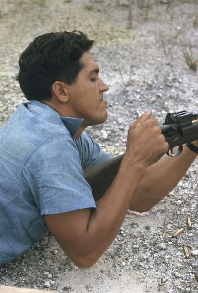 Rifle marksmanship class of the anti-Castro group Commandos L in the Everglades. A man is lying on the gravelly ground operating the bolt action of a rifle. A few spent cartridges are near him on the ground.