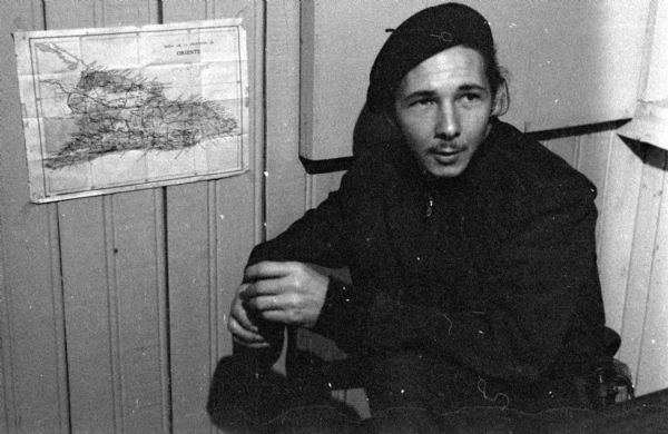 Raúl Castro sitting next to a small map of Oriente Province taped to the wall during the Cuban Revolution. The wall is bead-board and Castro is wearing a beret.