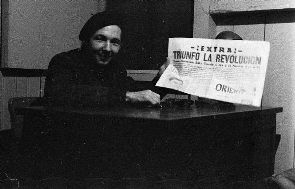 Raúl Castro sitting at a desk holding up a newspaper during the Cuban Revolution. The newspaper reads "!Extra! Triunfo La Revolution" and Castro is wearing a beret.
