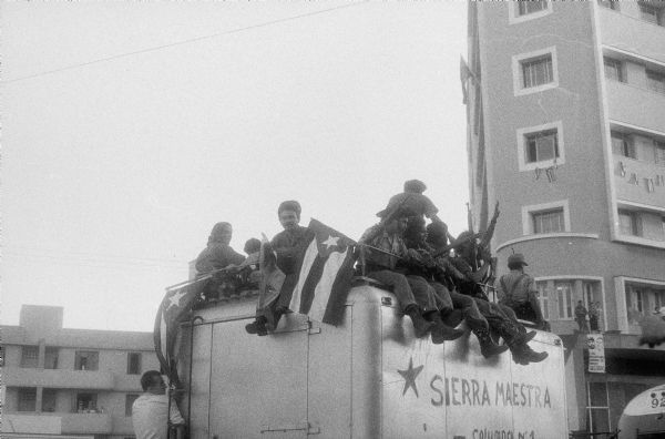 Parade in Havana to celebrate the success of the Cuban Revolution. A group of 26th of July Movement soldiers are sitting on top of a van as it is driving past apartment buildings. They are waving flags and rifles. The side of the van reads: "Sierra Maestra - Columna No. 1".