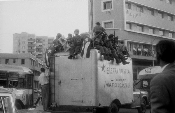Parade in Havana, Cuba celebrating the success of the Cuban Revolution. Soldiers holding flags, guns and a newspaper are riding on top of a truck. The side of the truck reads: "Sierra Maestra - Column No. 1 !Viva Fidel Castro!" There are apartment buildings in the background, other vehicles and onlookers are around the truck.