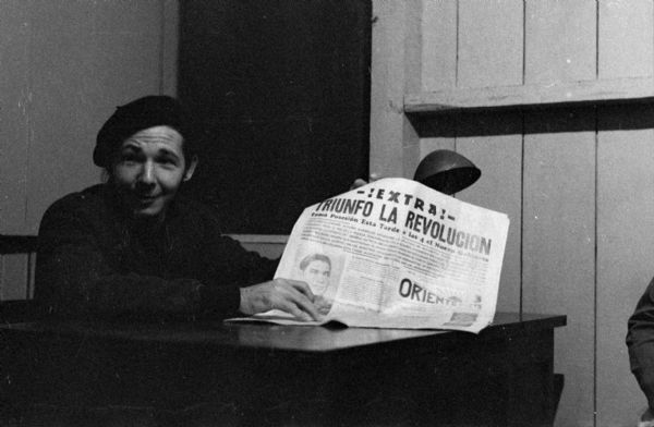 Raúl Castro sitting at a desk holding up a newspaper during the Cuban Revolution.  Castro is wearing a beret and the newspaper reads "!Extra! Triunfo La Revolution."