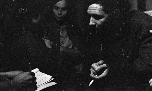 Raúl Castro is smoking a cigarette and leaning towards someone writing in a notebook during the Cuban Revolution. Vilma Espin and probably Celia Sanchez are talking in the background.