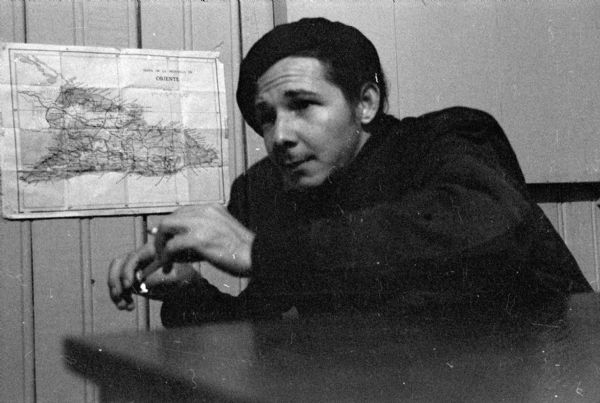 Raúl Castro sitting at a desk in front of a map of Oriente Province taped to the wall. He is wearing a beret and holding a cigarette in his hands.