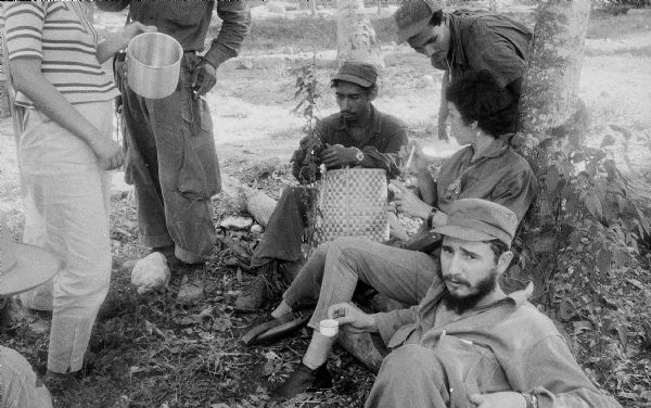 26th of July Movement rest by a tree in Oriente Province during the Cuban Revolution. Fidel Castro is holding an espresso cup in the foreground, and Celia Sánchez is leaning on the tree.